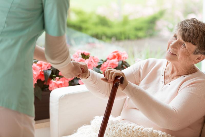 An older woman holds a cane and smiles at a nurse who is helping her stand up